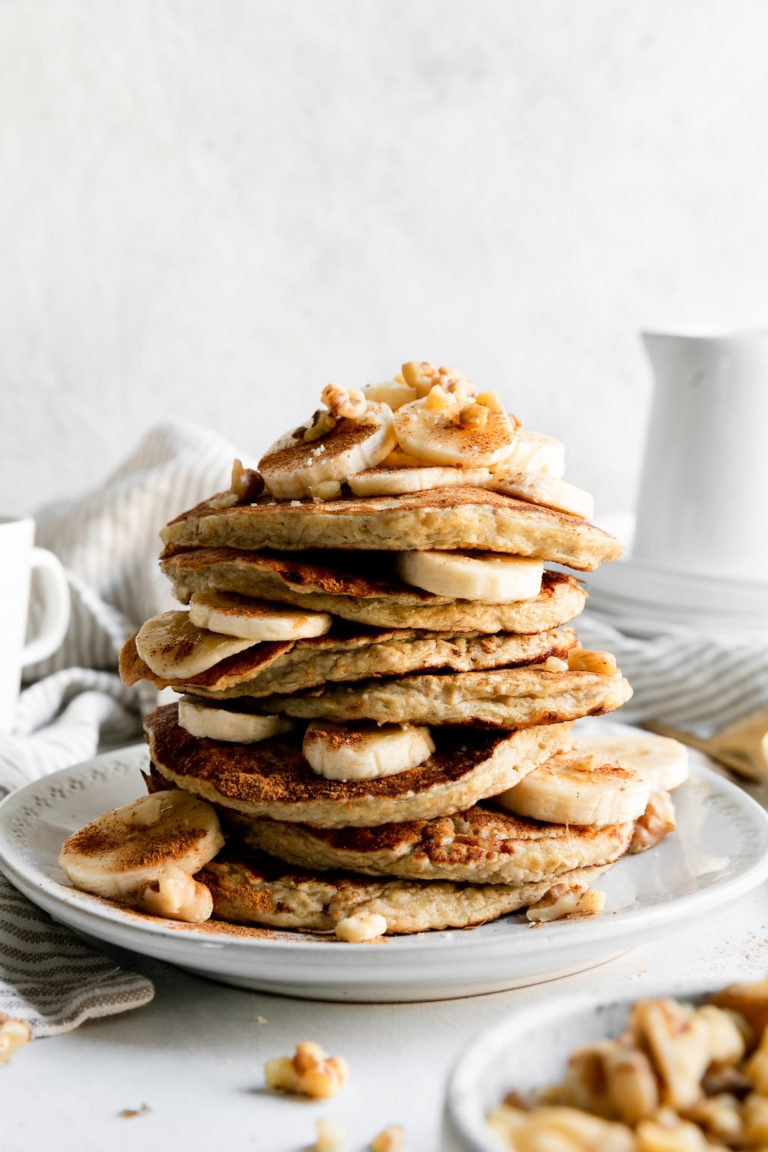 A tall stack of banana oatmeal pancakes with banana slices and walnuts stacked up on a white plate.