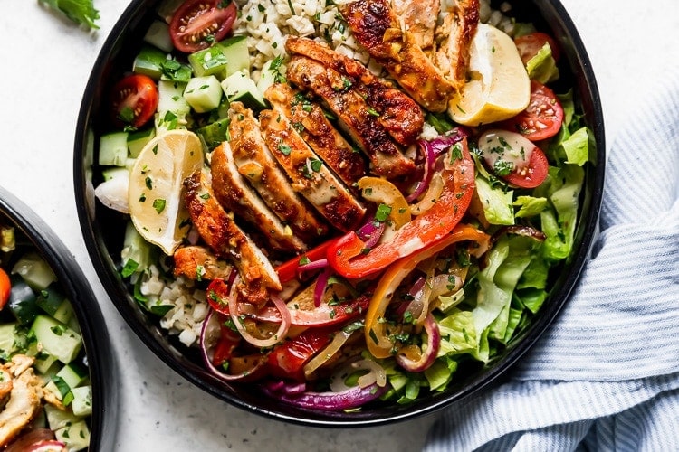 Sheet Pan Chicken Shawarma Bowls: A black bowl filled with cauliflower rice, roasted veggies, sliced chicken thighs, lettuce, cucumbers, and cherry tomatoes and drizzled with a cilantro-lime sauce.