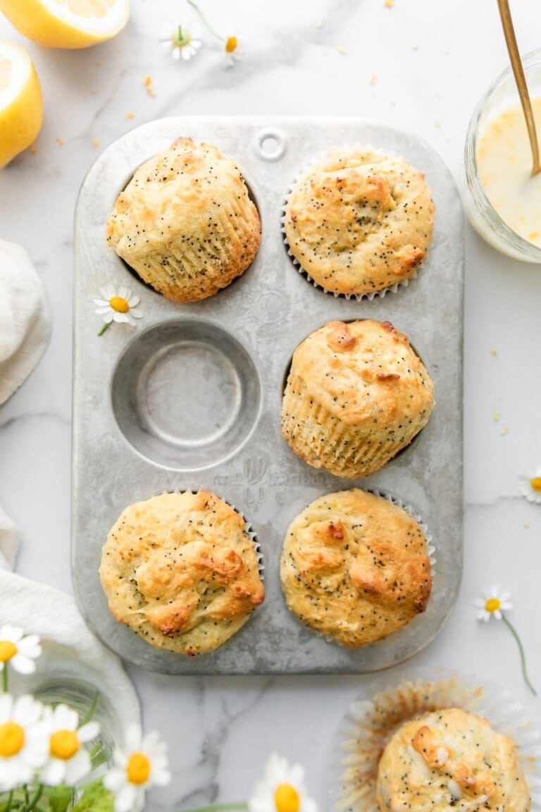 A silver six count muffin tin filled with five lemon poppyseed muffins, one muffin on the counter topped with lemon glaze.
