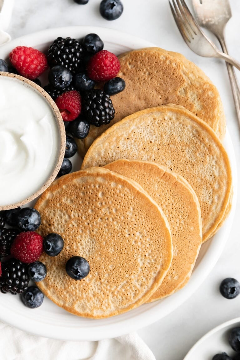Several protein pancakes on white plate with fresh berries and small bowl of yogurt